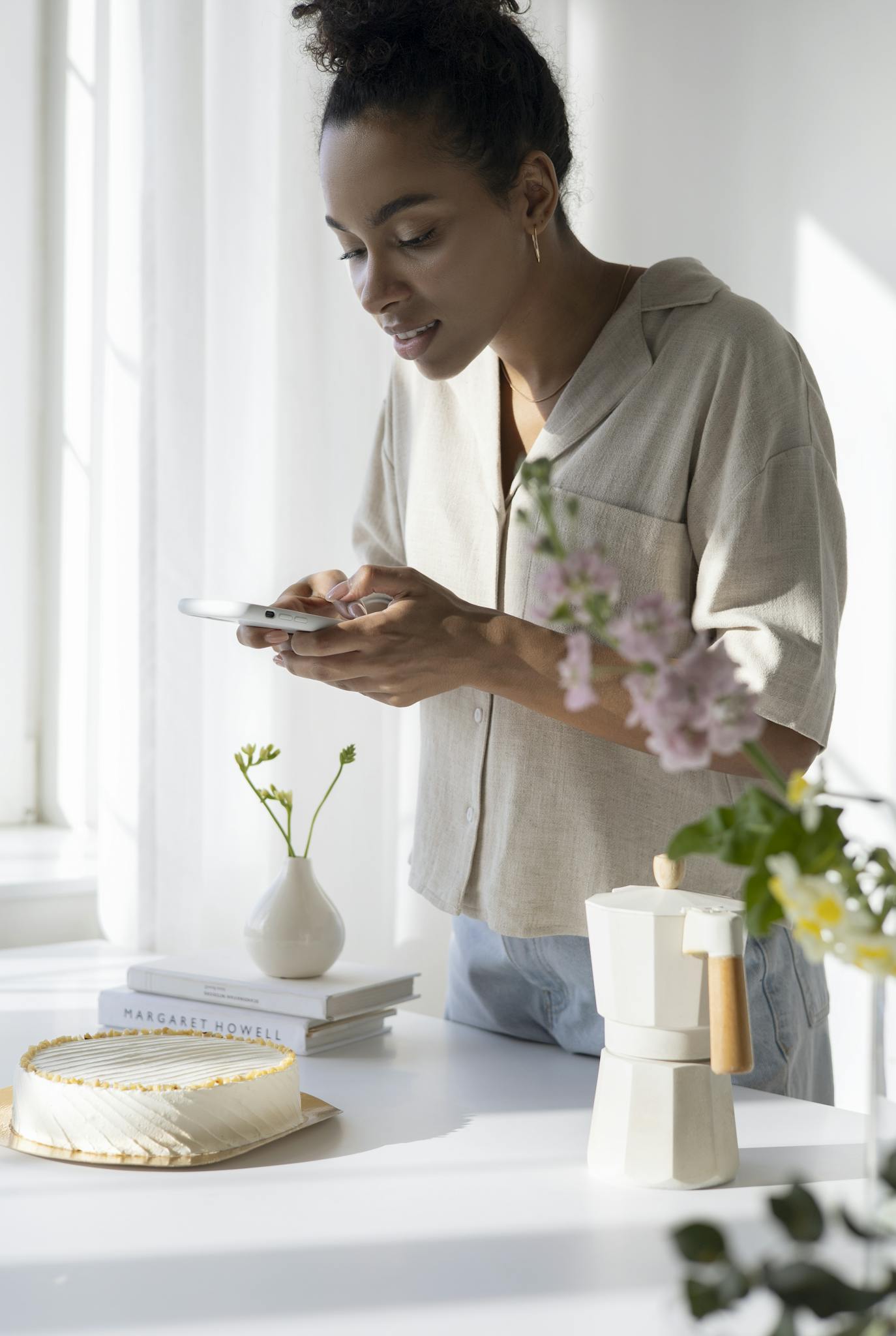 Woman capturing a stylish coffee setup in a sunlit kitchen with flowers and cake.